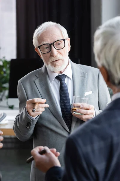 Empresário idoso em óculos segurando charuto e copo de uísque perto colega em primeiro plano borrado — Fotografia de Stock
