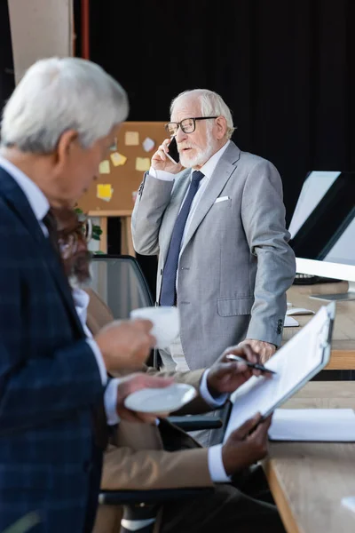 Senior businessman in eyeglasses talking on mobile phone near blurred interracial colleagues — Stock Photo