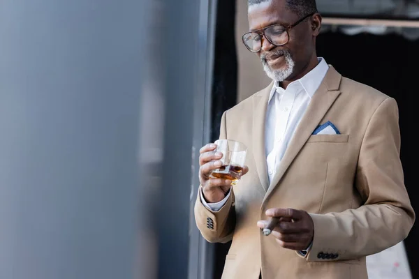 Exitoso empresario afroamericano de pie con cigarro y vaso de whisky en la oficina - foto de stock