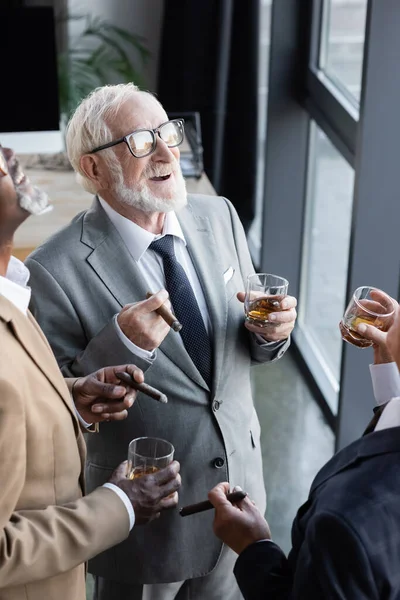 Vue en grand angle des principaux partenaires d'affaires multiethniques avec des cigares et des verres de whisky riant au bureau — Photo de stock