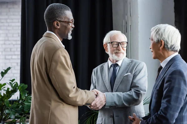 Glückliche interrassische Geschäftspartner beim Händeschütteln in der Nähe eines älteren asiatischen Kollegen — Stockfoto