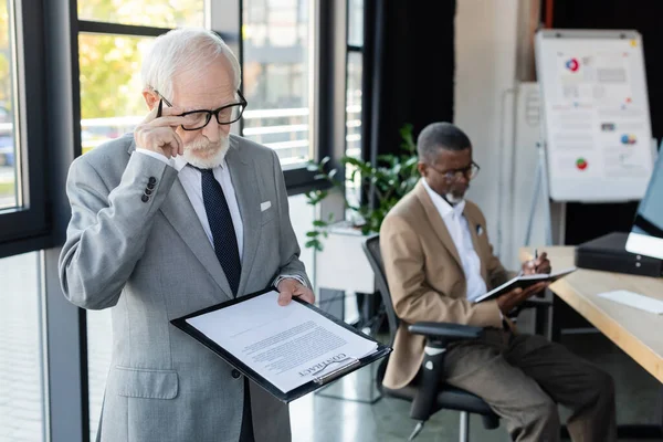 Старший бізнесмен читає контракт, в той час як афроамериканський колега працює на розмитому фоні — стокове фото