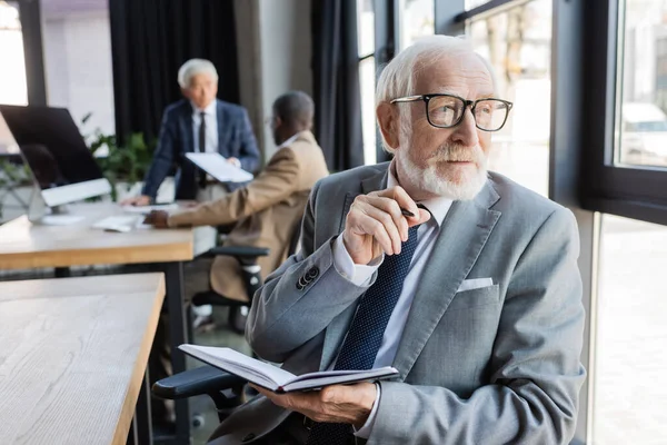 Senior businessman holding notebook tandis que des collègues multiethniques travaillent sur fond flou — Stock Photo