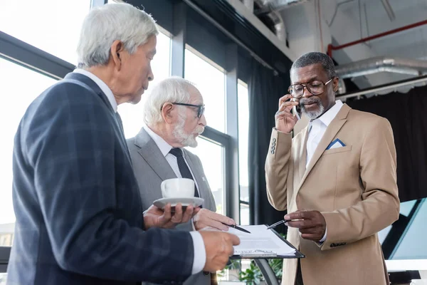 Американський бізнесмен-афроамериканець розмовляє на смартфоні поруч з контрактом і старшими колегами — стокове фото