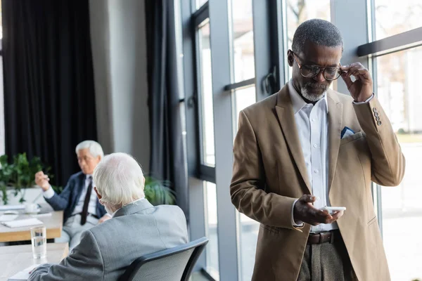 Anziano uomo d'affari afroamericano che regola gli occhiali mentre guarda smartphone vicino a colleghi sfocati — Foto stock