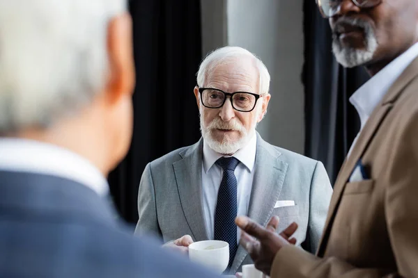Elderly businessman in eyeglasses holding coffee cup near blurred interracial colleagues — Stock Photo
