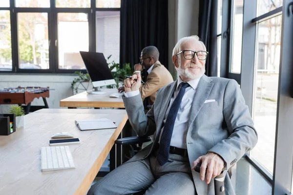 Feliz hombre de negocios sénior mirando a través de la ventana mientras que el colega afroamericano que trabaja en fondo borroso - foto de stock