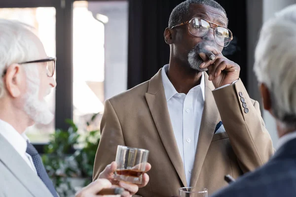 Homme d'affaires afro-américain senior en lunettes fumant un cigare près de collègues flous avec des verres de whisky — Photo de stock