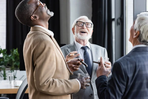 Hombres de negocios multiétnicos mayores riendo mientras beben whisky y fuman cigarros en la oficina - foto de stock
