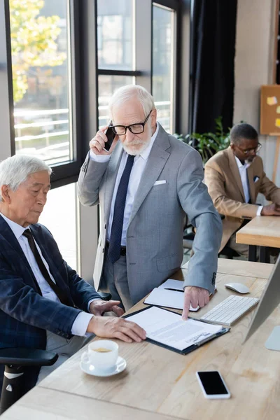 Senior businessman pointing at contract near asian colleague while talking on mobile phone — Stock Photo