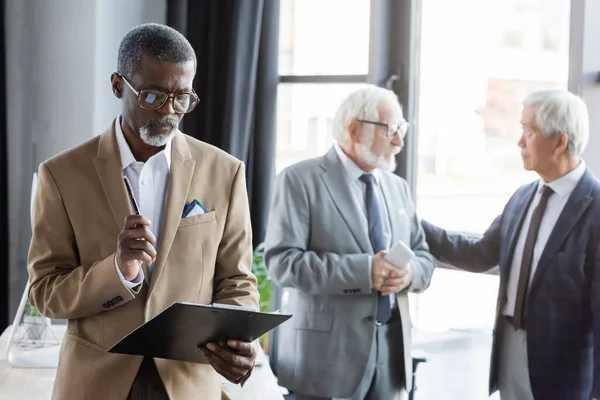 Старший африканский американский бизнесмен смотрит в буфер обмена рядом со старшими коллегами, разговаривая на размытом фоне — стоковое фото