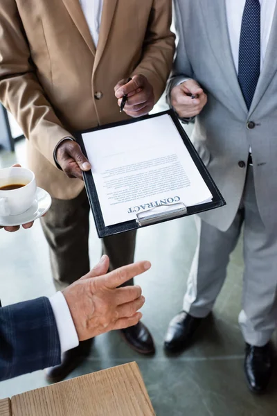 Cropped view of senior businessman with cup of coffee near interracial colleagues with contract on clipboard — Stock Photo