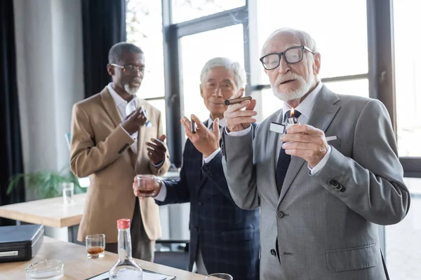 Respetable hombre de negocios de alto nivel sosteniendo encendedor y cigarro cerca borrosa colegas interracial con vasos de whisky - foto de stock