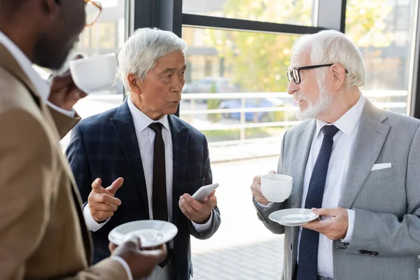 Sênior asiático empresário com smartphone apontando com o dedo enquanto conversa com colegas bebendo café — Fotografia de Stock