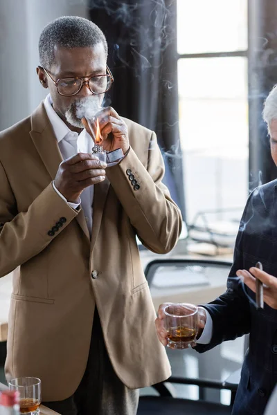 Hombre de negocios afroamericano en anteojos iluminación cigarro cerca colega con vaso de whisky - foto de stock