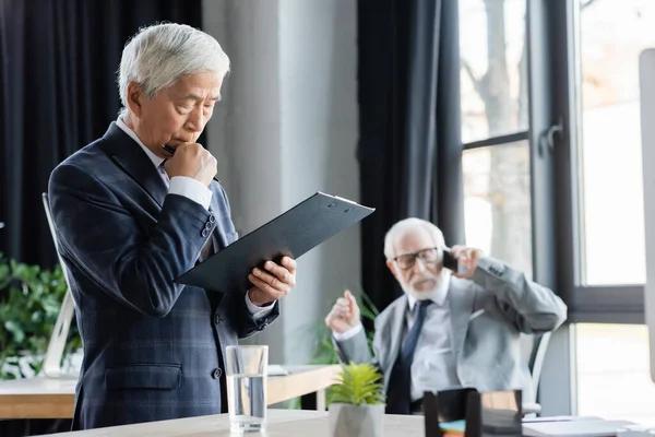 Pensativo asiático empresário olhando para prancheta perto sênior colega falando no smartphone no fundo borrado — Fotografia de Stock