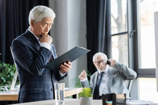 Thoughtful asian businessman looking at clipboard near senior colleague talking on smartphone on blurred background — Stock Photo