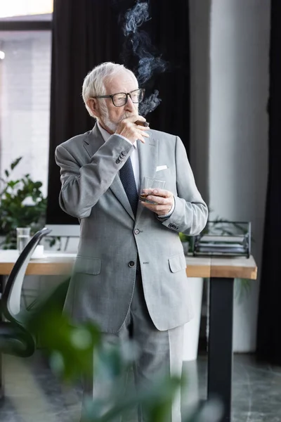 Anziano uomo d'affari in giacca e cravatta fumare sigaro mentre tiene un bicchiere di whisky — Foto stock