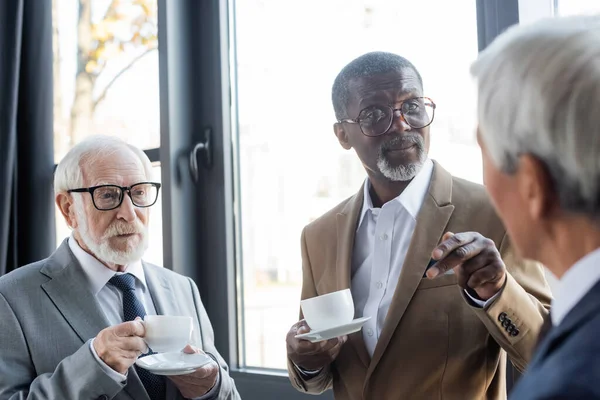 Літні багатонаціональні бізнесмени з чашками кави дивиться на колегу на розмитому передньому плані — стокове фото