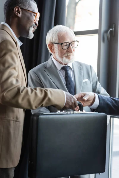 Senior interracial hommes d'affaires donnant mallette à partenaire d'affaires dans le bureau — Photo de stock