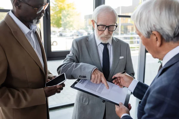 Старший бизнесмен указывает на контракт рядом с межрасовыми коллегами с ручкой и смартфоном — стоковое фото