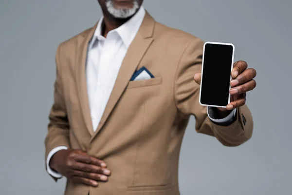 Vista recortada de un empresario afroamericano borroso mostrando un teléfono inteligente con pantalla en blanco aislado en gris - foto de stock
