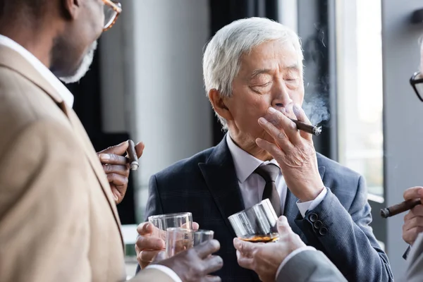 Senior asian businessman smoking cigar while clinking glasses of whiskey with blurred interracial colleagues — Stock Photo