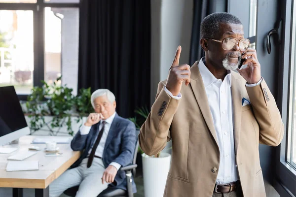 African american businessman pointing with finger while talking on smartphone near blurred asian colleague — Stock Photo
