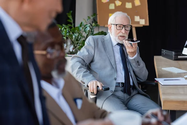 Senior businessman sending voice message on smartphone near blurred multiethnic colleagues — Stock Photo