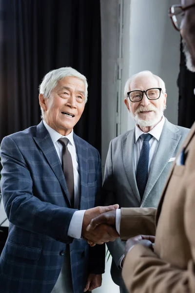 Веселий азіатський бізнесмен тремтить руками розмитого афроамериканця біля старшого колеги — стокове фото