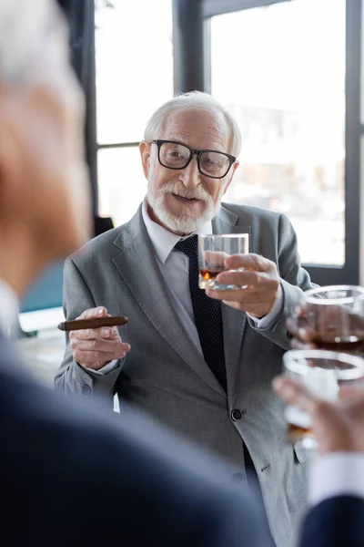 Homme d'affaires senior avec cigare et verre de whisky parler à un collègue flou dans le bureau — Photo de stock