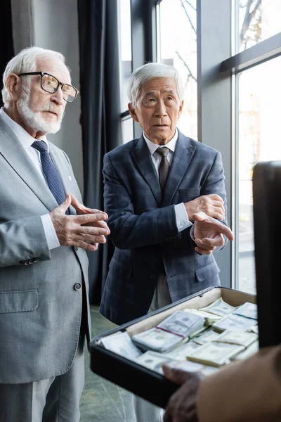 Asiático hombre de negocios apuntando con el dedo en maletín con dinero cerca senior colega - foto de stock