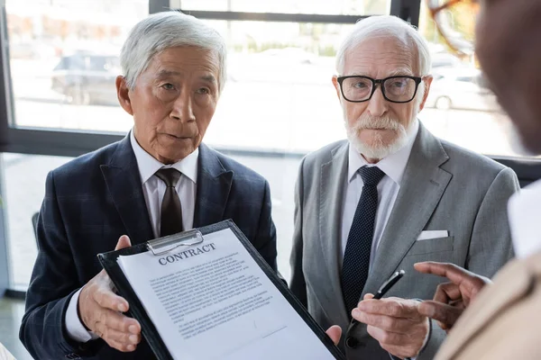 Colegas interracial senior celebración de contrato y pluma cerca borrosa socio de negocios afroamericano - foto de stock