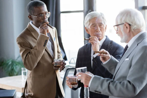 Elderly multiethnic business people with cigars and glasses of whiskey talking in office — Stock Photo