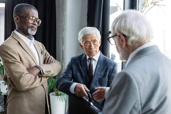 Старший азиатский бизнесмен дарит портфель размытому деловому партнеру рядом с африканским американцем со скрещенными руками — стоковое фото