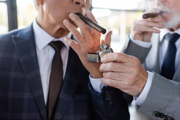 Cropped view of blurred senior businessman lighting cigar of business partner in office — Stock Photo