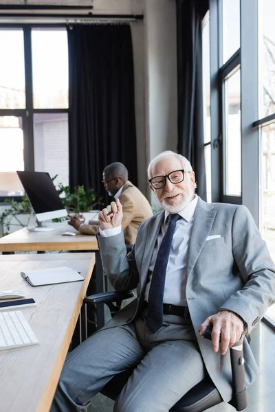 Felice uomo d'affari anziano sorridente alla macchina fotografica mentre collega afro-americano che lavora su sfondo sfocato — Foto stock