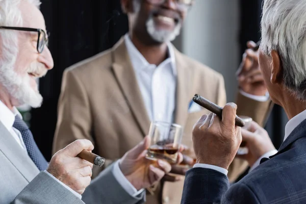 Hombre de negocios senior sosteniendo un vaso de whisky y cigarro cerca de colegas multiétnicos sonrientes - foto de stock