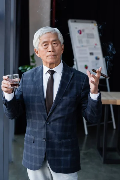 Senior et confiant asiatique homme d'affaires avec cigare et verre de whisky regardant caméra dans le bureau — Photo de stock