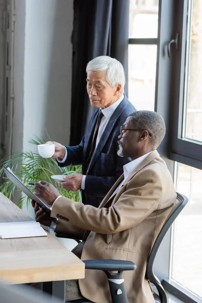 African american businessman showing document to senior asian colleague with cup of coffee — Stock Photo
