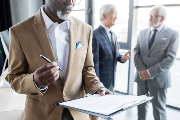 Afrikanischer amerikanischer Geschäftsmann hält Stift und Vertrag in der Nähe hochrangiger Kollegen, die über verschwommenen Hintergrund sprechen — Stockfoto