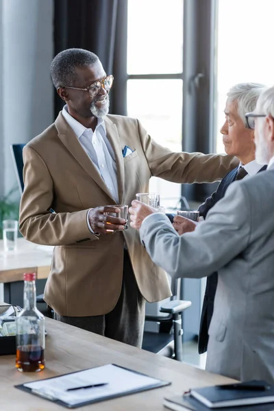 Feliz africano americano hombre de negocios tintineo vasos con multiétnicos colegas cerca de contrato en escritorio - foto de stock