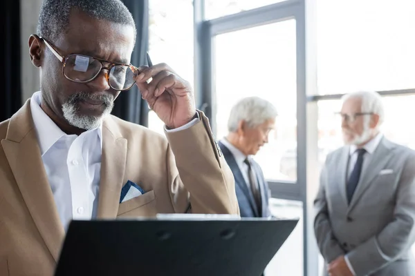 Hombre de negocios afroamericano senior con portapapeles tocando anteojos mientras sus colegas hablan sobre un fondo borroso - foto de stock