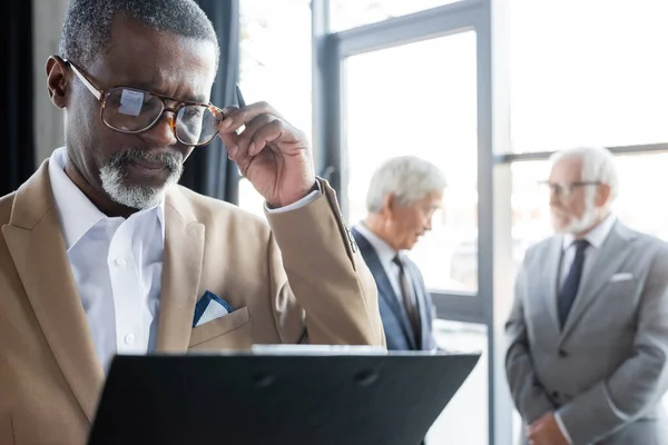 Senior afrikanisch-amerikanischer Geschäftsmann mit Klemmbrett, das Brillen berührt, während Kollegen auf verschwommenem Hintergrund sprechen — Stockfoto