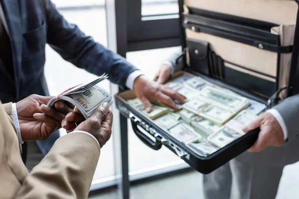 Vista recortada do empresário afro-americano segurando notas de dólar perto de colegas borrados e maleta com dinheiro — Fotografia de Stock