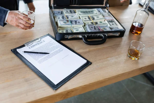 Vue partielle de l'homme d'affaires senior près du contrat, mallette avec des dollars et du whisky sur le bureau — Photo de stock