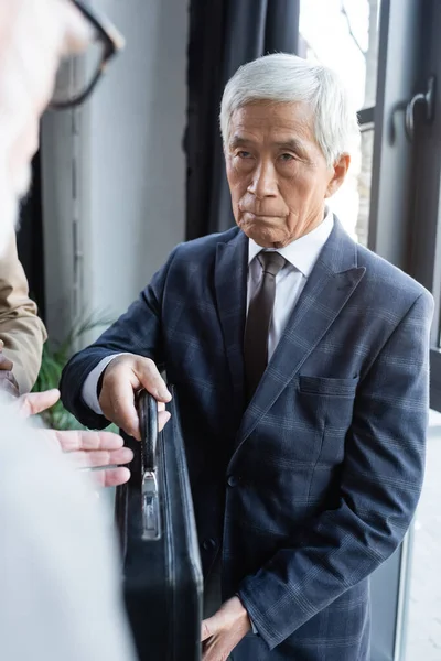 Senior und selbstbewusster asiatischer Geschäftsmann gibt Aktentasche an verschwommenen Geschäftspartner — Stockfoto