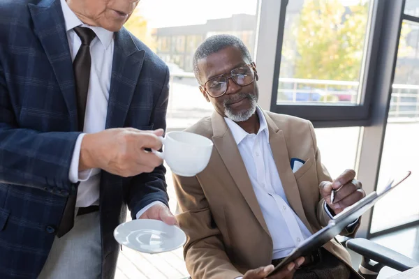 Empresário americano africano sênior apontando com caneta na área de transferência perto colega com xícara de café — Fotografia de Stock