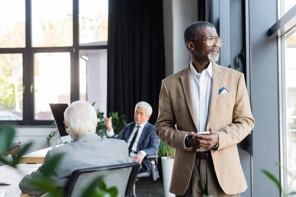 Homme d'affaires afro-américain avec smartphone debout près de la fenêtre tandis que des collègues flous travaillant dans le bureau — Photo de stock