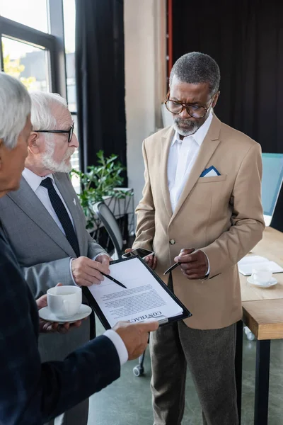 Hombre mayor señalando con pluma en el contrato cerca de socio de negocios afroamericano y colega con taza de café - foto de stock